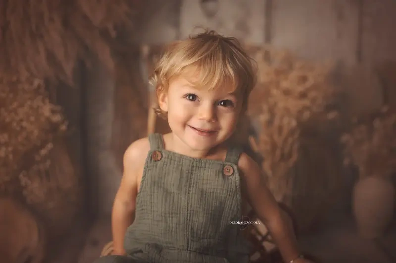 Blond toddler in khaki green gauze dungarees, sitting and smiling in front of a backdrop of golden dried flowers